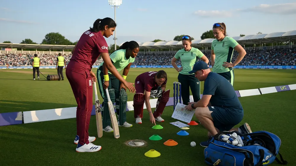 Women cricketers in maroon and mint kits gathered with a coach around multicoloured cones and a tactics sheet on a green outfield